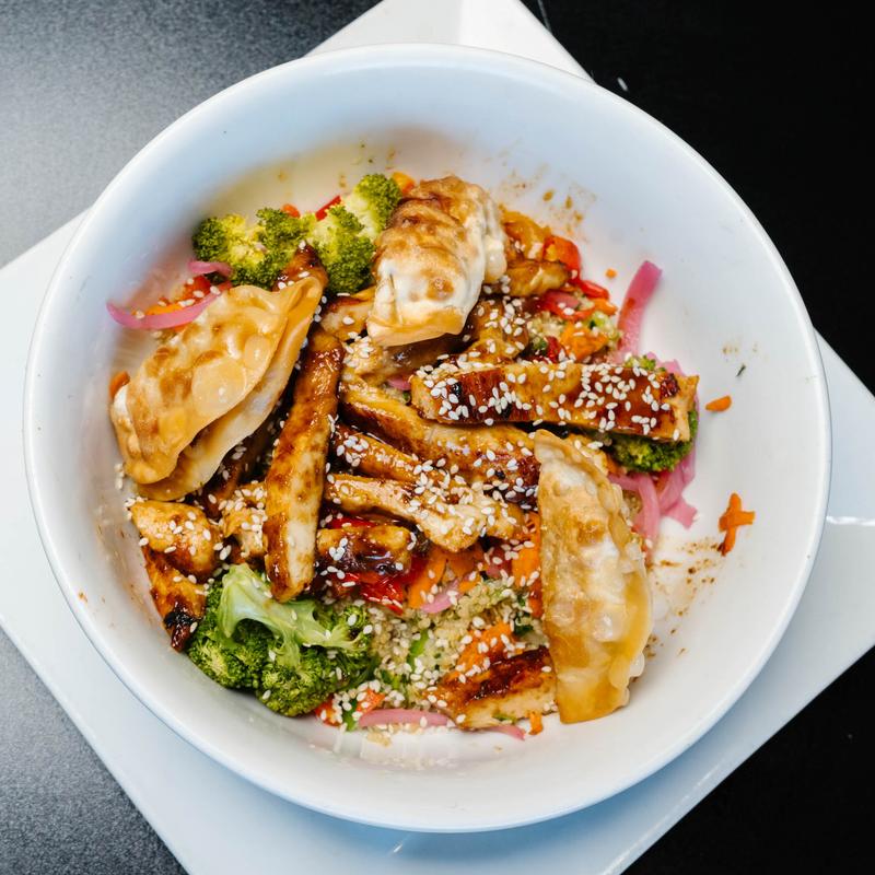 White bowl with grilled tofu strips topped with sesame seeds, pieces of broccoli, pickled red onions, shredded carrots, and three crispy potstickers, placed on a white square plate on a black surface.