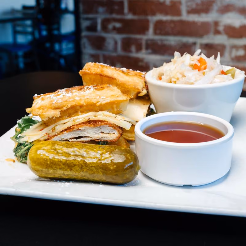 Plate with a sliced sandwich containing bread, cheese, and greens, a whole pickle, a white bowl of coleslaw, and a small white bowl of dipping sauce.