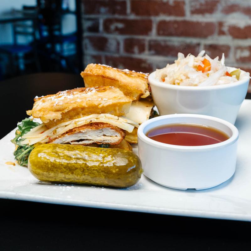 Plate with a sliced sandwich containing bread, cheese, and greens, a whole pickle, a white bowl of coleslaw, and a small white bowl of dipping sauce.
