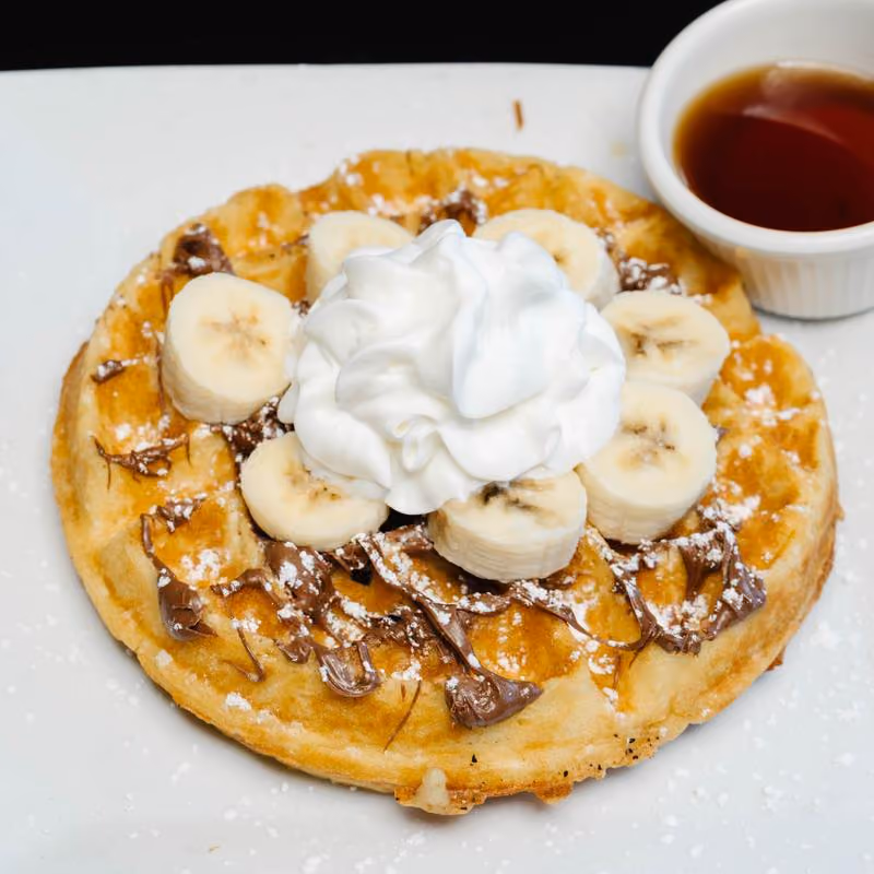 Waffle topped with sliced bananas, dollop of whipped cream, and drizzled with chocolate spread on a white plate next to a small cup of syrup.