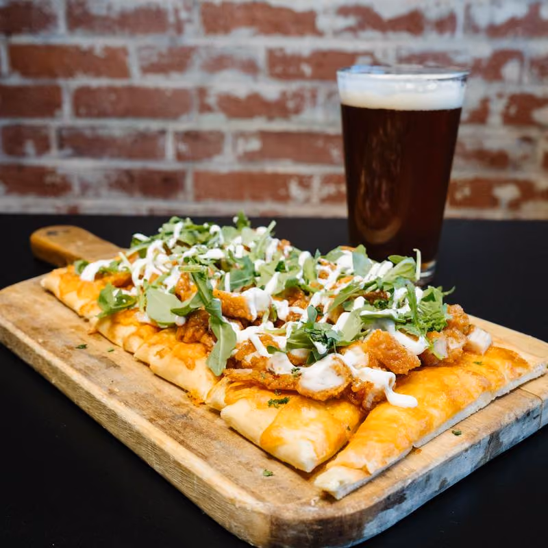 Flatbread pizza with melted cheese, crispy chicken pieces, fresh greens, and drizzled white sauce on a wooden board, next to a glass of dark beer with foam on top, set against a brick wall background.