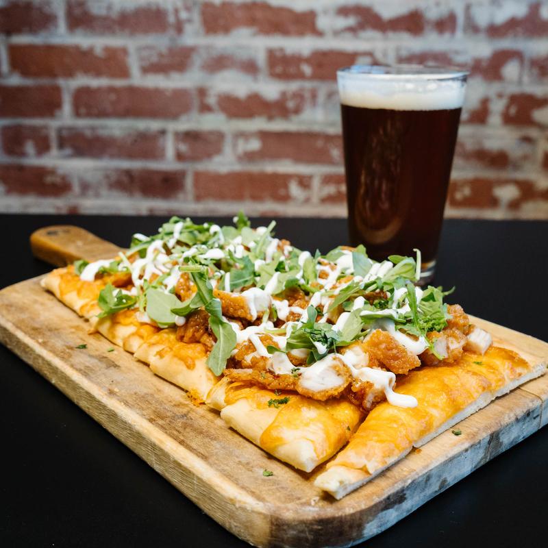 Flatbread pizza with melted cheese, crispy chicken pieces, fresh greens, and drizzled white sauce on a wooden board, next to a glass of dark beer with foam on top, set against a brick wall background.