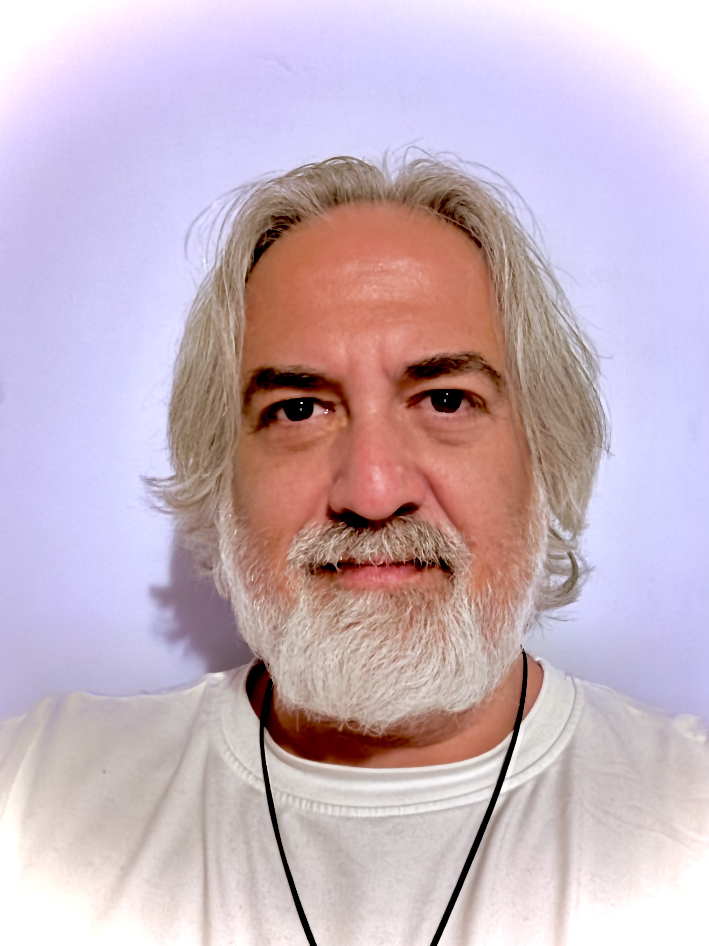 Portrait of a middle-aged man with medium-length gray hair and a full white beard wearing a white shirt and a black cord necklace, against a plain pale background.