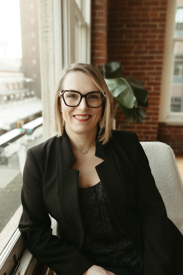 Kathleen Fugere, ARNP, seated in a black blazer beside a bright window, smiling, with an exposed brick wall and a large leafy plant behind her