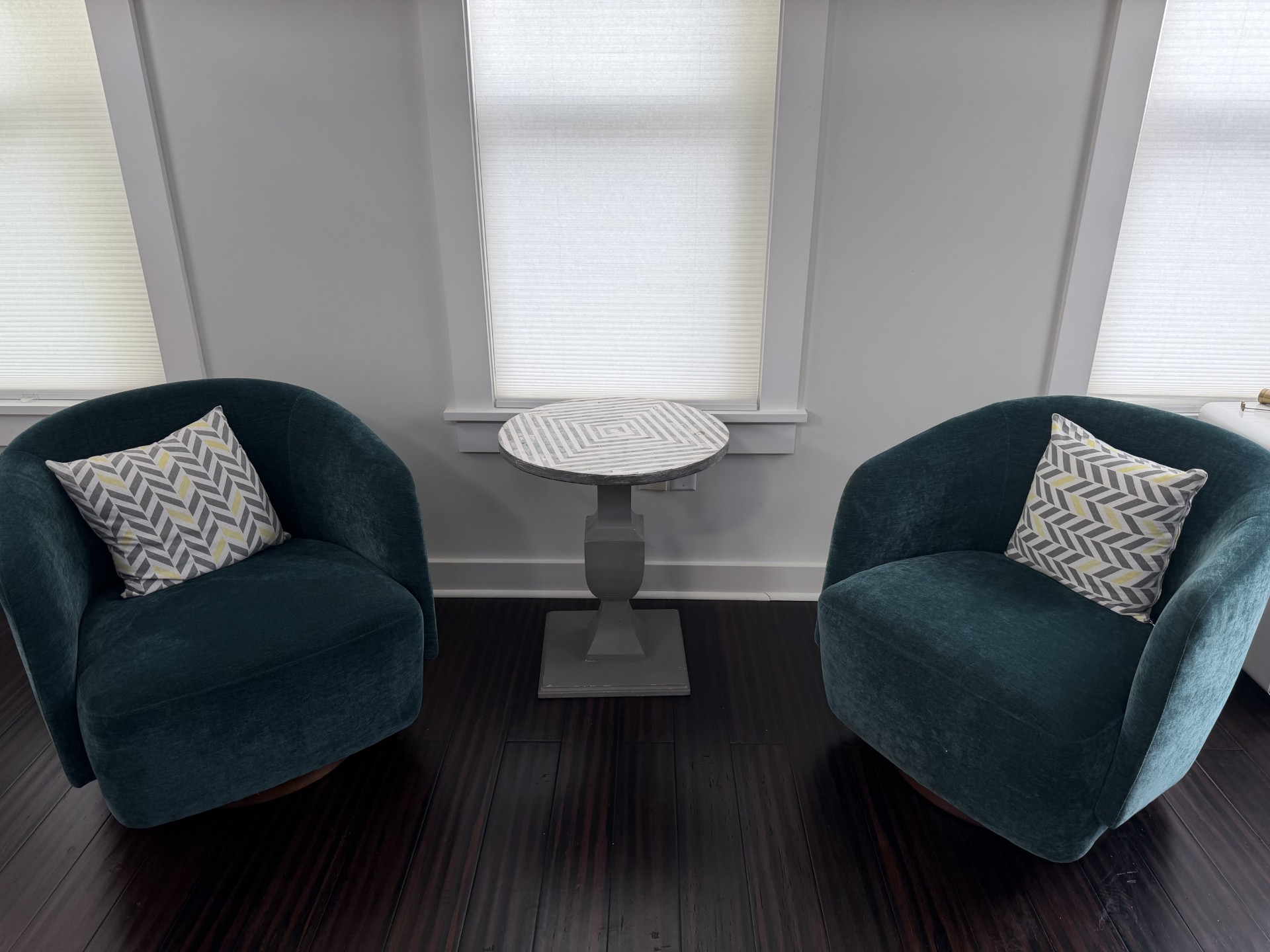 Two teal velvet armchairs facing a small round side table beside tall office windows with light filtering through the shades