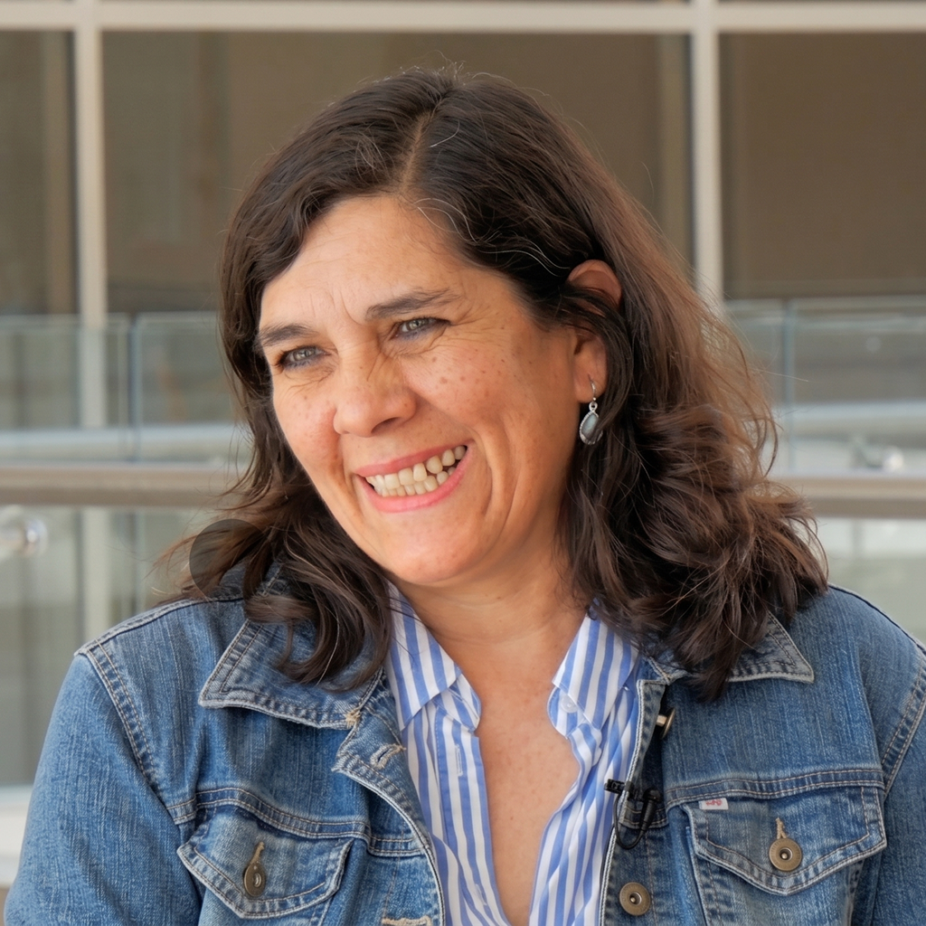 Mujer sonriente con cabello castaño oscuro y rizado, viste chaqueta de mezclilla azul y camisa con rayas blancas y azules, con aretes plateados, en un entorno interior con baranda transparente al fondo.