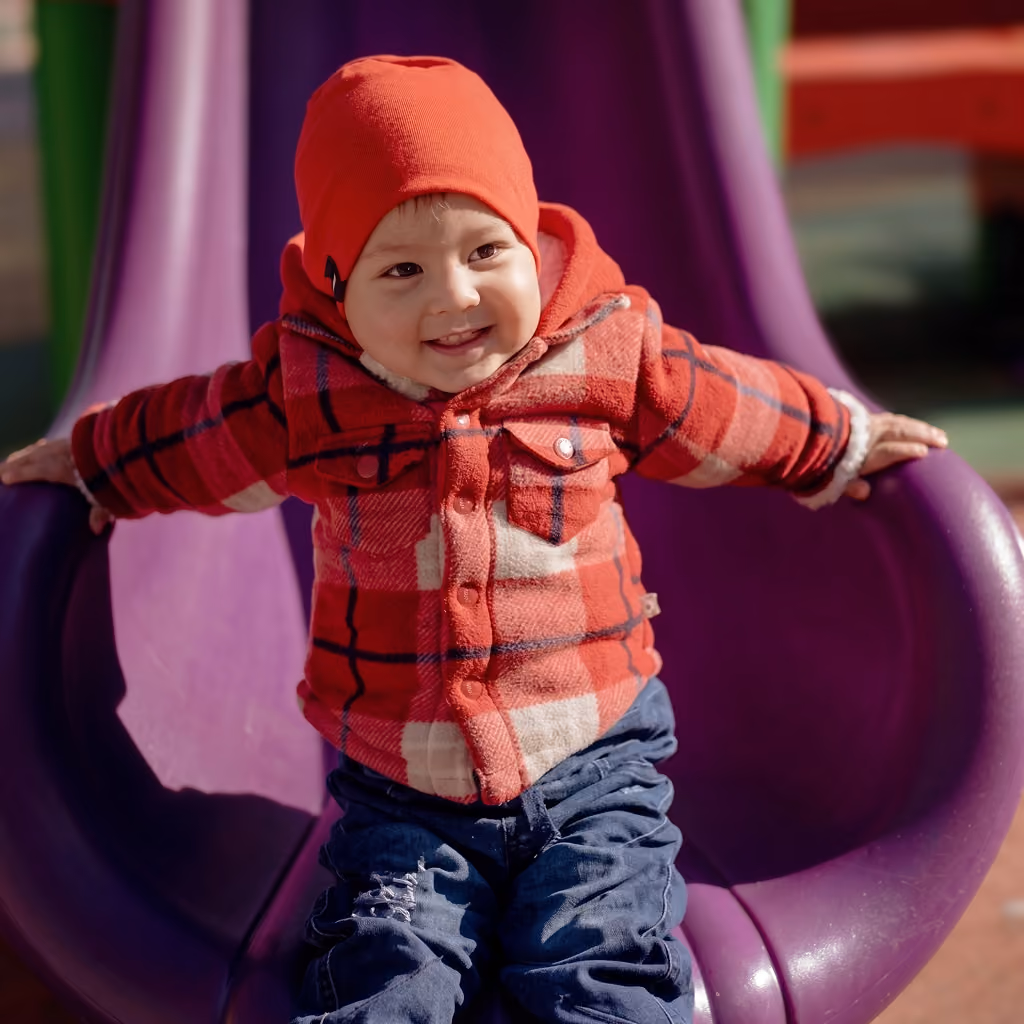Niño pequeño sonriente con gorro rojo y chaqueta a cuadros rojos y blancos sentado en un tobogán morado en un parque.