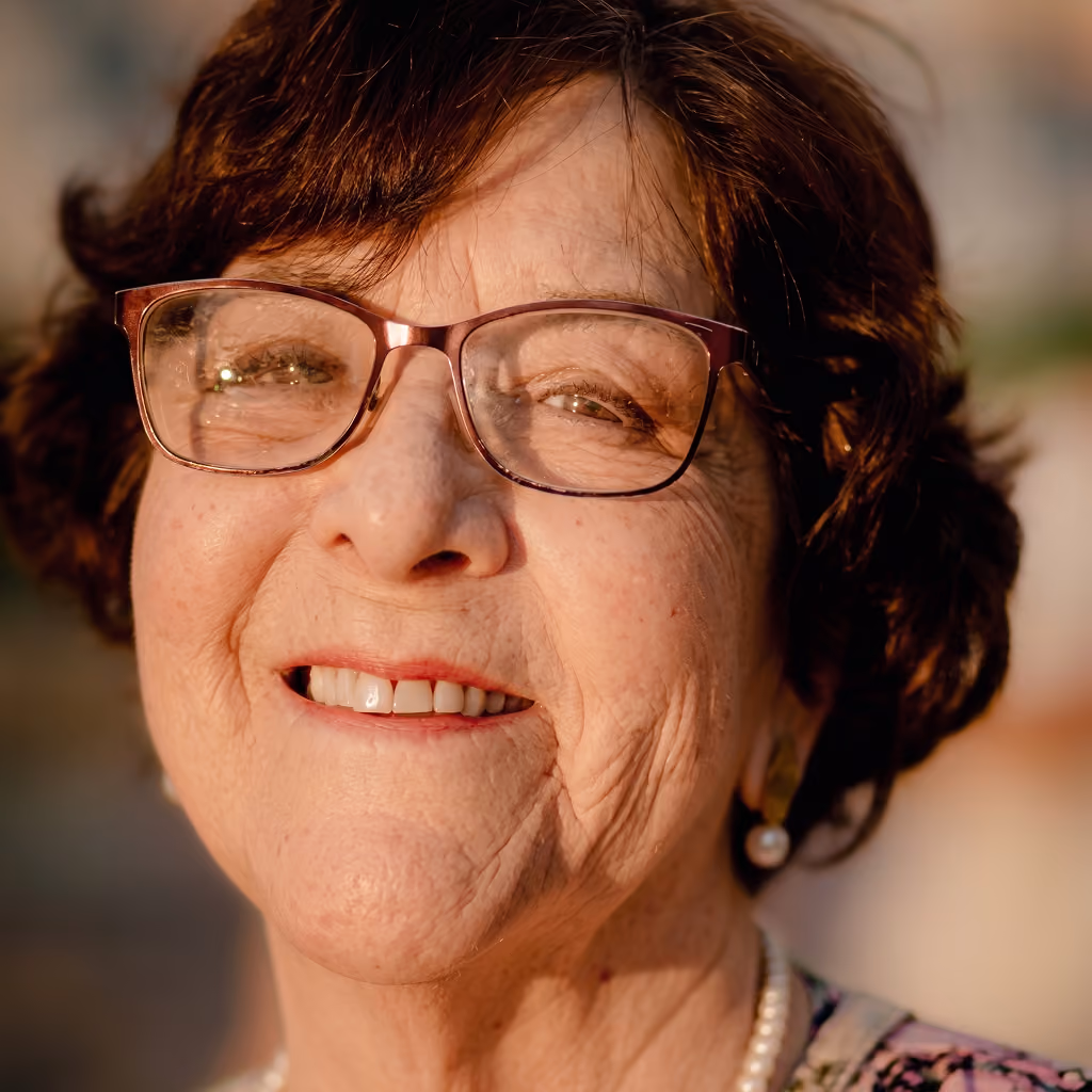 Primer plano de una mujer mayor con gafas, cabello corto y oscuro, sonriendo con una expresión amable y relajada, lleva un collar de perlas y un pendiente de perla.