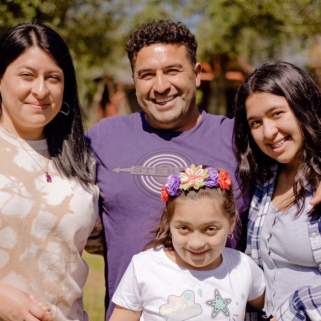 Fotografía de un grupo familiar de cuatro personas sonriendo al aire libre, con dos mujeres adultas, un hombre y una niña con una diadema floral colorida.
