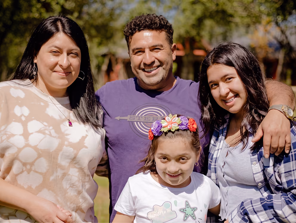 Cuatro personas sonrientes al aire libre en un día soleado: un hombre con camiseta morada y tres mujeres, una niña con diadema de flores, una mujer joven con camisa a cuadros y una mujer con camiseta beige y blanca.