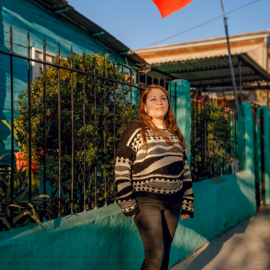 Mujer de cabello castaño claro con suéter blanco y negro de diseño geométrico, de pie frente a una reja de hierro y una casa pintada de turquesa bajo luz de sol al atardecer.