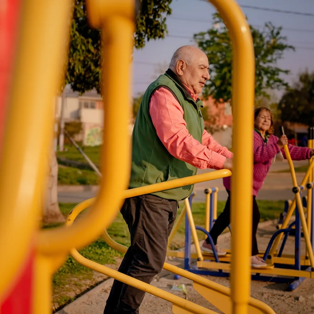 Hombre mayor con chaleco verde y camisa rosada utilizando una máquina de ejercicio al aire libre en un parque con estructuras amarillas, mientras una mujer mayor con suéter morado hace ejercicio en el fondo.