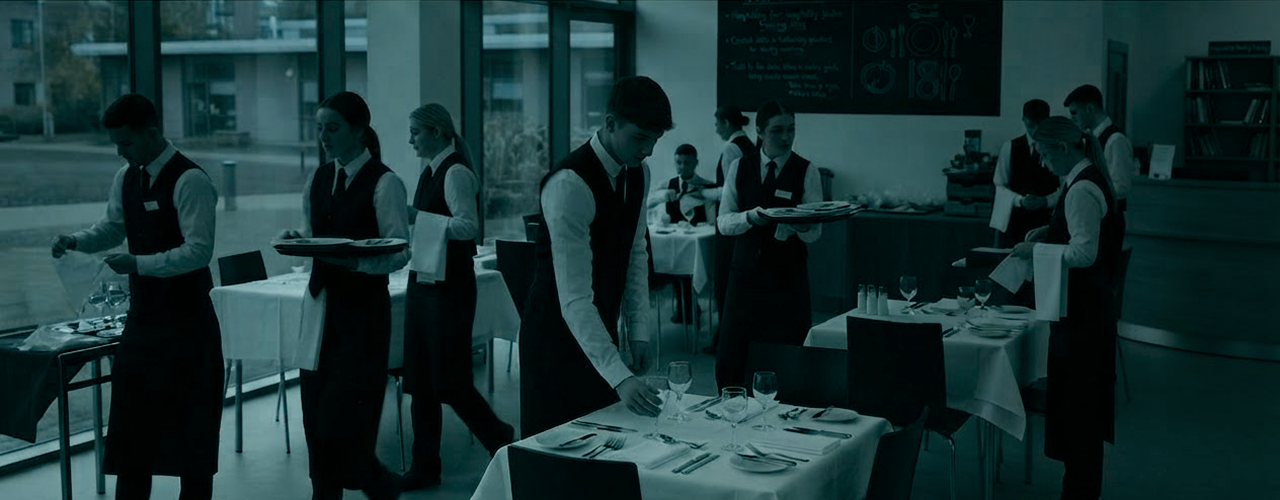 A group of servers in black vests and white shirts preparing tables in a restaurant, arranging glasses and plates on white tablecloths near large windows.