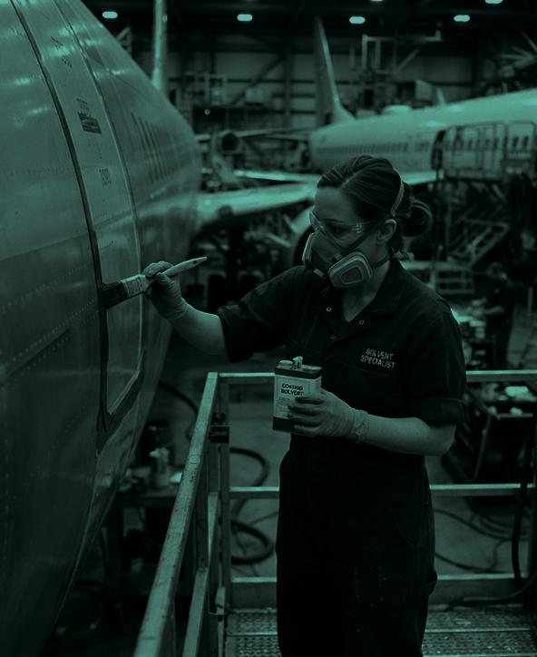 Person wearing safety goggles, a respirator mask, gloves, and a jumpsuit labelled 'Solvent Specialist' applying solvent with a brush to the side of an aircraft inside a large hangar with other planes present.