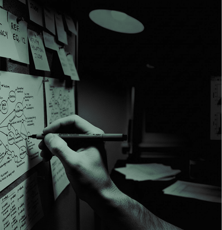 A hand holding a marker pen writing or drawing diagrams and notes on sticky notes and paper pinned to a board, in a dimly lit room.