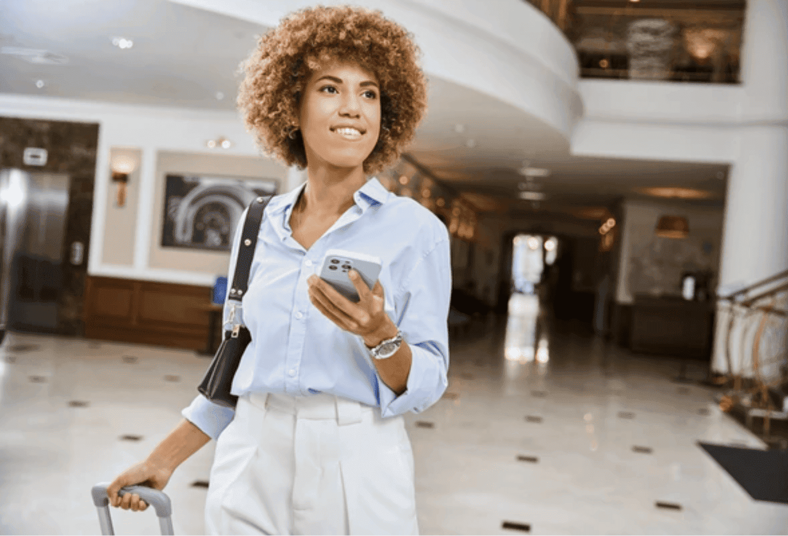 Smiling woman with curly hair pulling suitcase and holding phone in a hotel lobby.