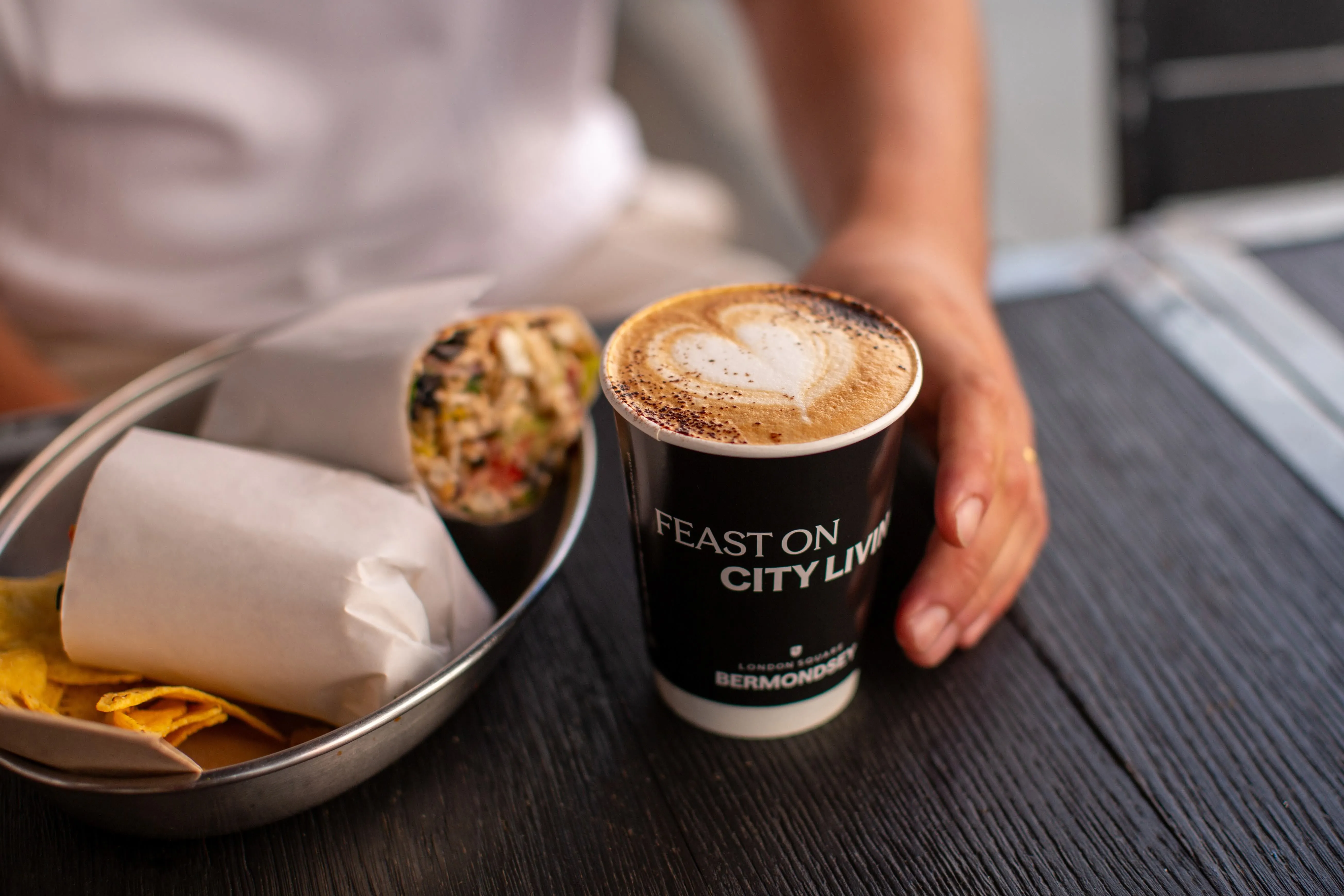 Black 'Feast on city living' branded takeaway coffee cup beside a burrito on an outdoor café table