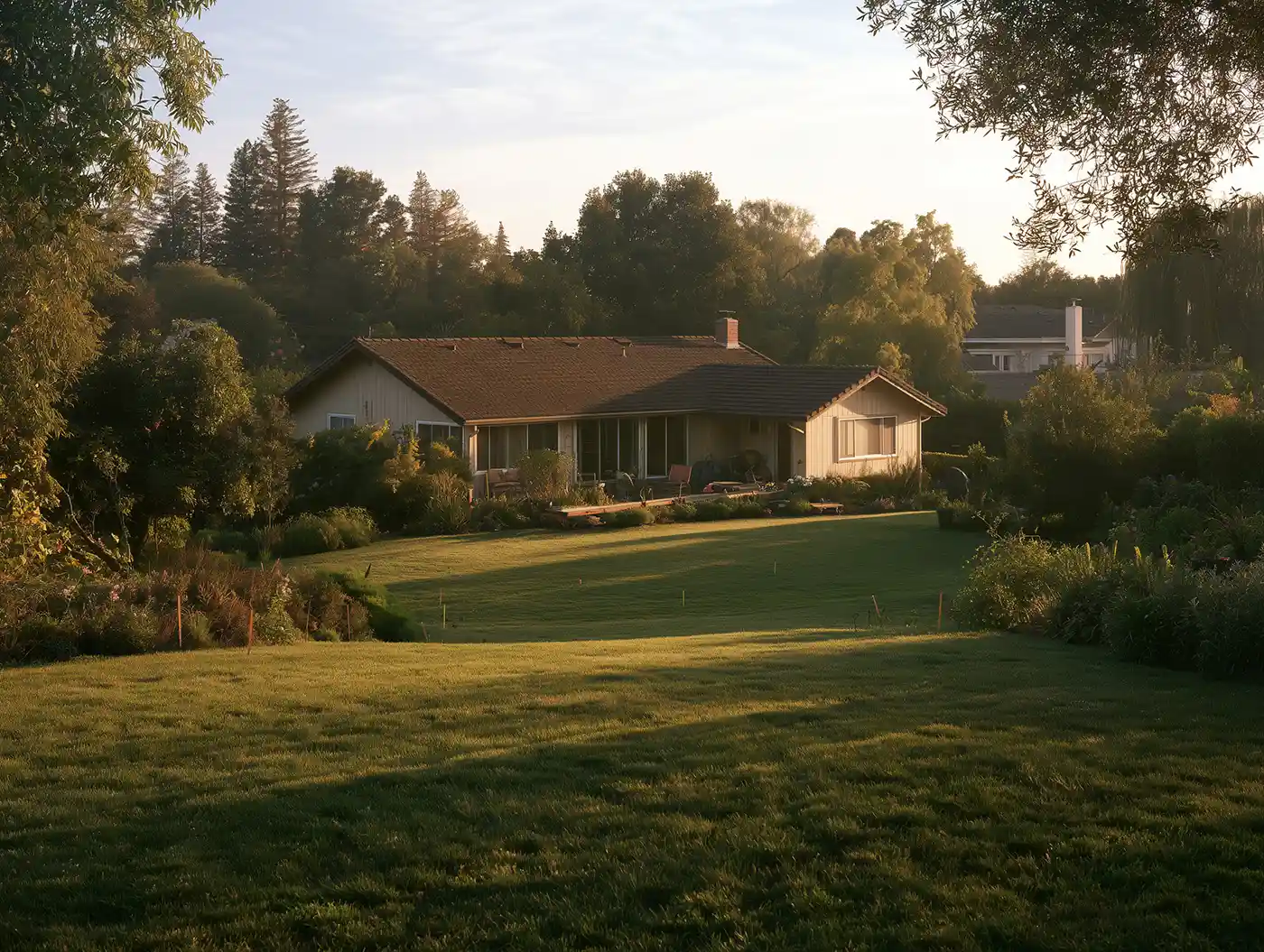 A single-story home nestled among green trees and a sloping lawn in the warm evening light.
