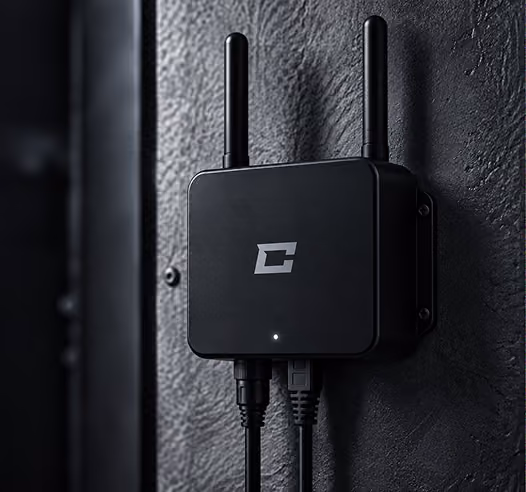 Black wireless router with two antennas mounted on a textured dark wall, with two cables connected at the bottom and a small white indicator light on the front.