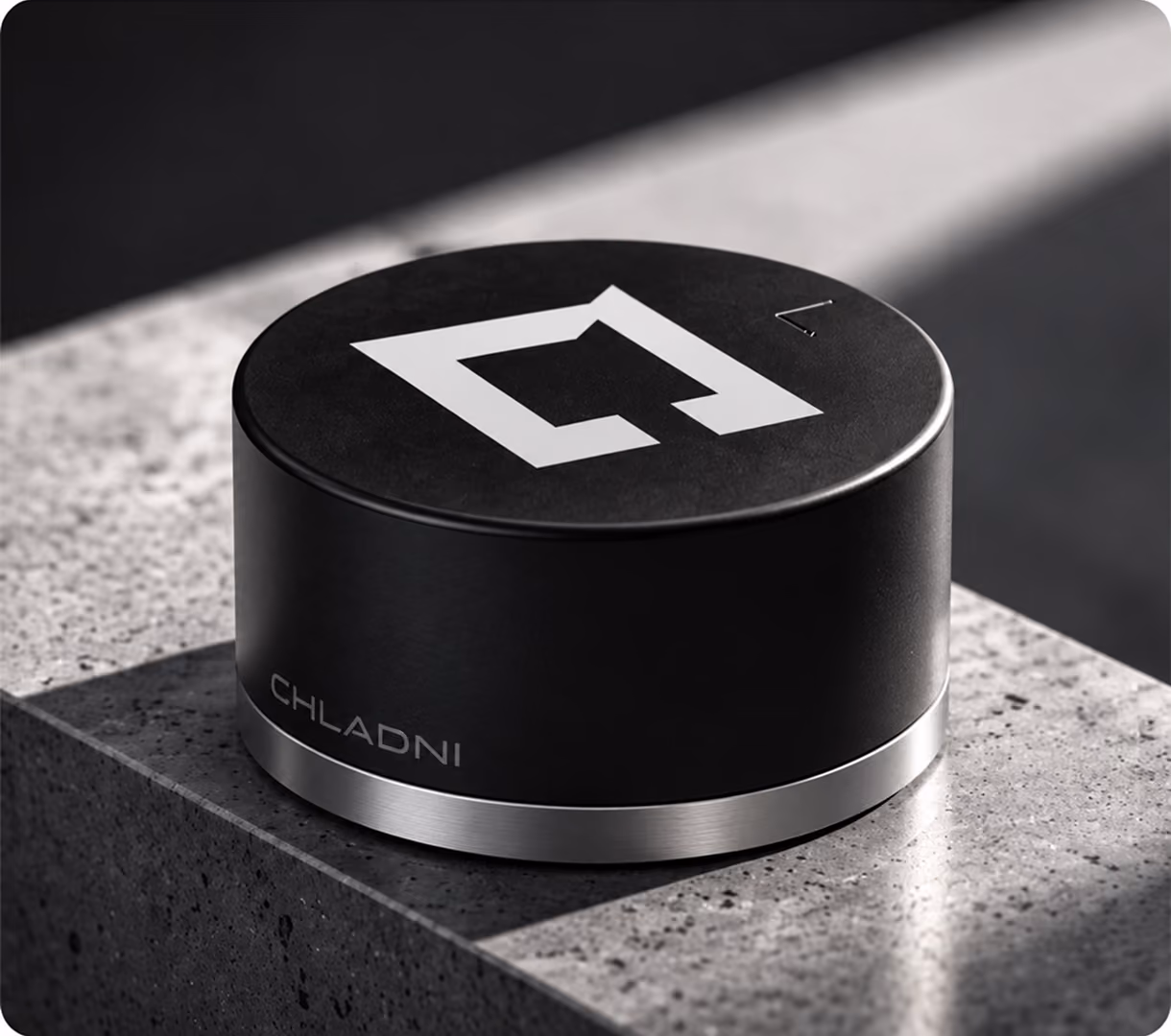 Black cylindrical device with a silver base and a white geometric logo on top, placed on a textured gray stone surface with sunlight casting shadows.