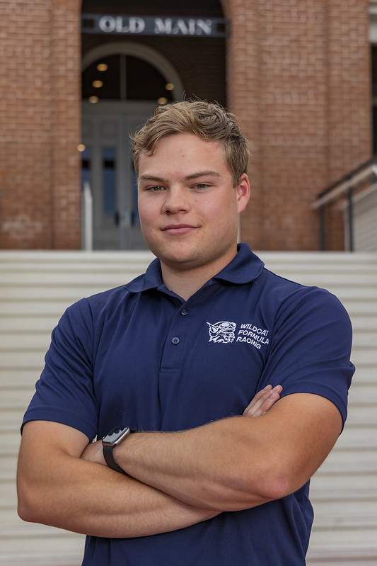 Grant Hastings with crossed arms wearing a navy blue polo shirt with a Wildcat Formula Racing logo, standing confidently in front of brick steps and the "Old Main" building on the University of Arizona campus.