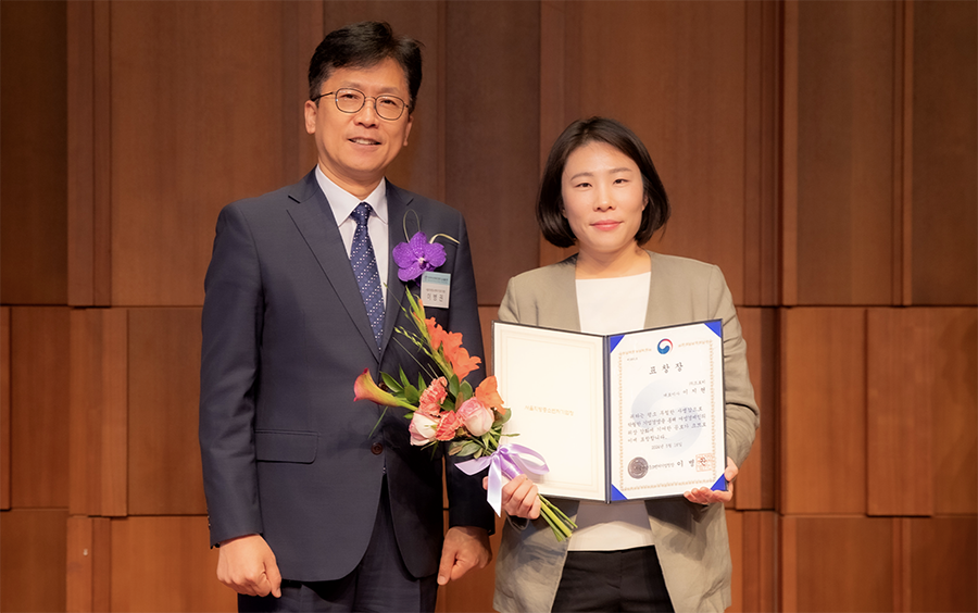 A man in a navy suit and tie stands next to a woman holding a bouquet of flowers and an award certificate in a blue folder, both smiling in front of a wooden panel backdrop.