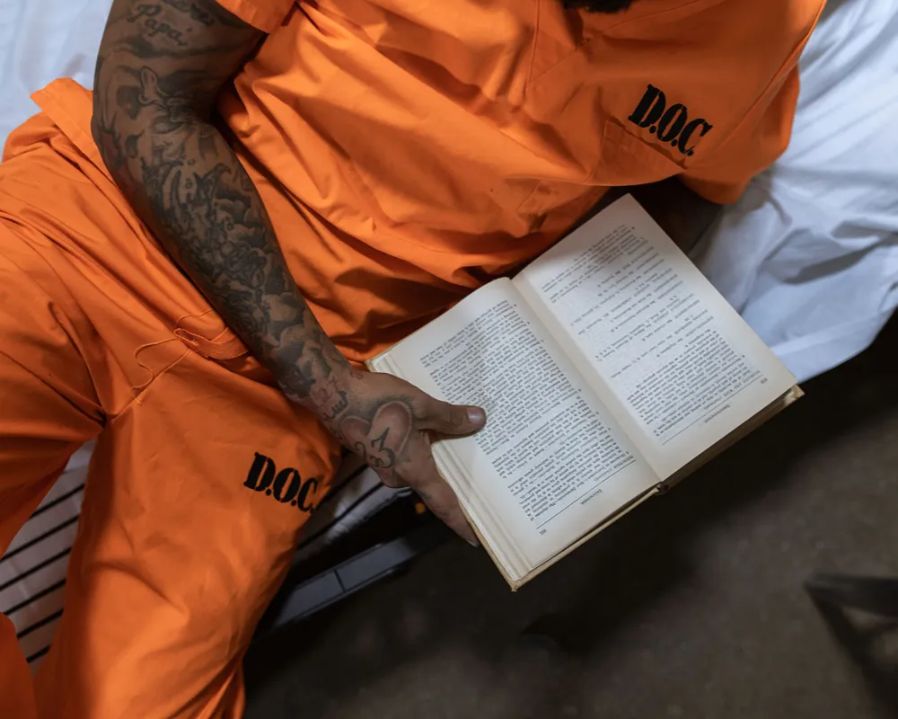 A person in an orange Department of Corrections uniform reads a Bible in a jail cell