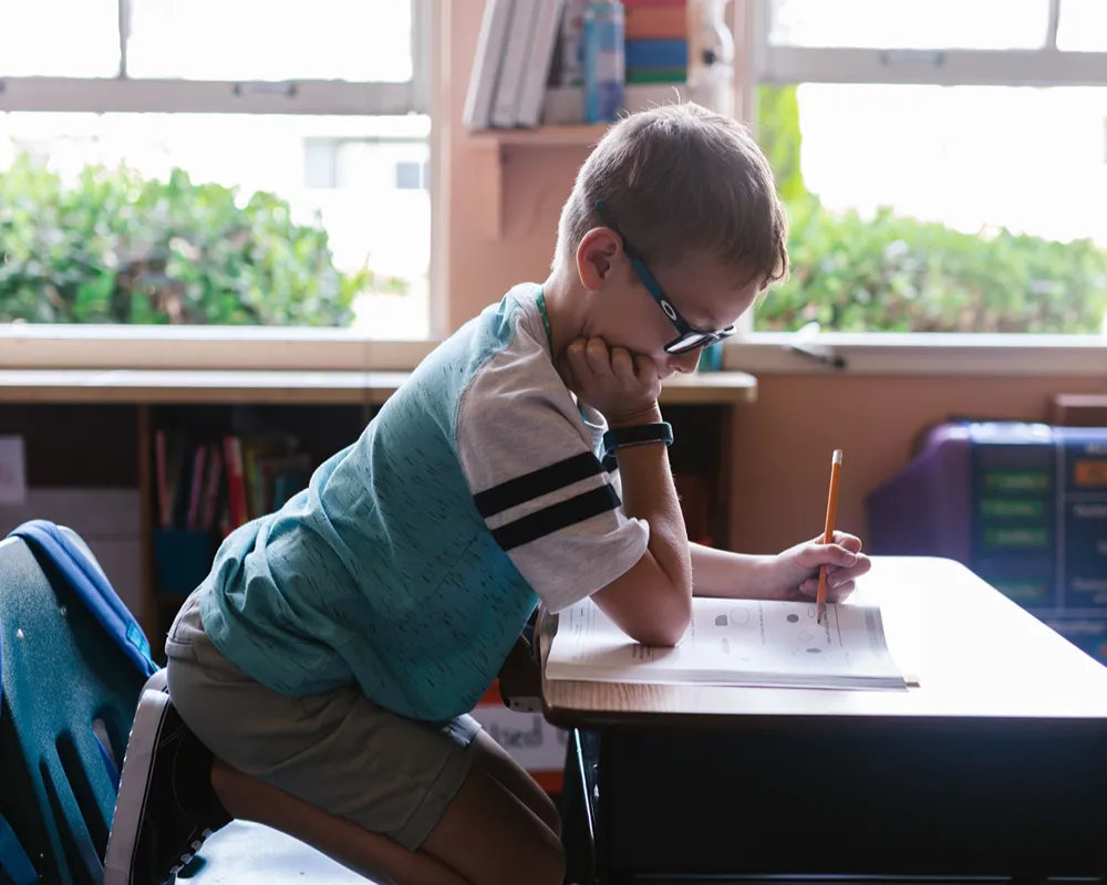 A young boy in glasses concentrates while writing with a pencil at a classroom desk