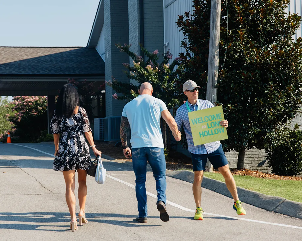 A volunteer holds a Welcome to Long Hollow sign as a couple walks toward the church entrance