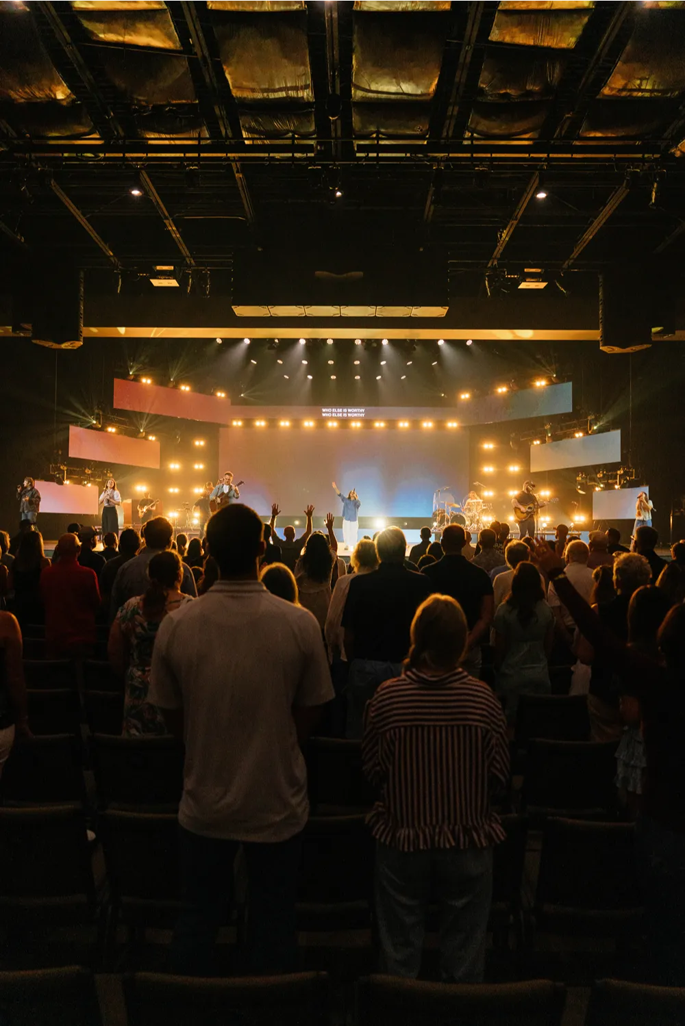 The congregation stands during worship as the band leads from a brightly lit stage