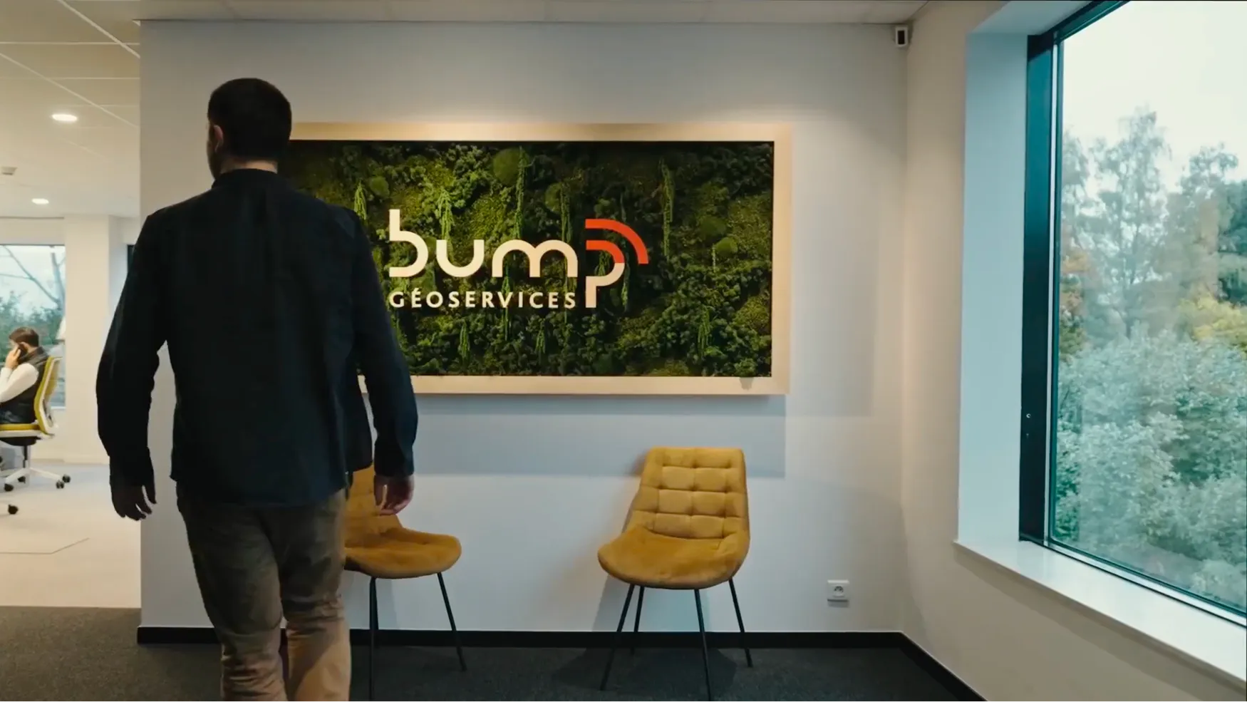 Un homme de dos marche dans un bureau moderne avec deux chaises jaunes et un panneau mural végétalisé affichant le logo de Bump Géoservices à côté d'une grande fenêtre donnant sur des arbres verts.