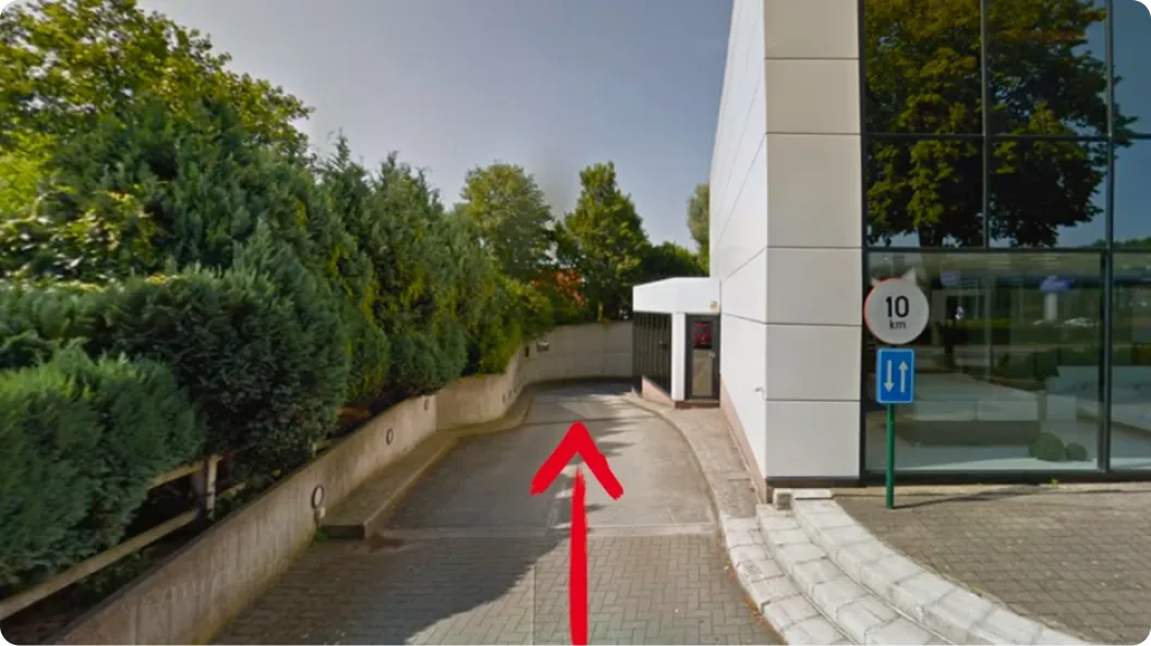 Entrée d'un parking avec une route en descente bordée de haies côté gauche et un bâtiment moderne à droite avec une façade vitrée, un panneau indiquant une vitesse maximale de 10 km/h et un panneau de circulation indiquant une voie bidirectionnelle. Une flèche rouge peinte au sol pointe vers l'entrée.