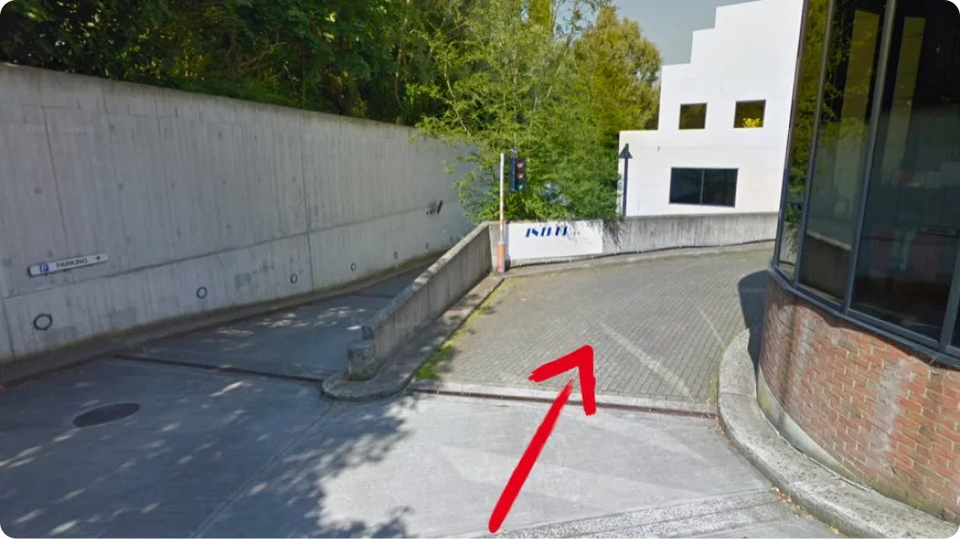Extérieur d'un chemin pavé avec une flèche rouge peinte au sol indiquant vers le haut et à droite, un bâtiment blanc à fenêtres carrées et une structure en briques avec de grandes fenêtres sur la droite.