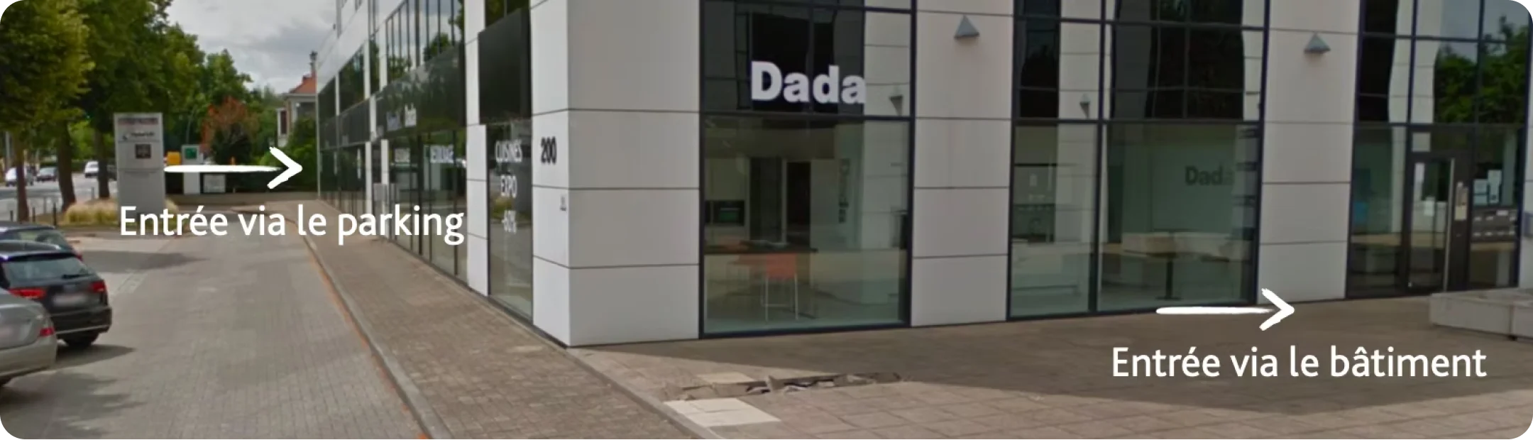 Façade d'un bâtiment moderne avec vitrines, au coin d'une rue pavée, indiquant deux entrées : une flèche vers la gauche pour l'entrée via le parking et une flèche vers la droite pour l'entrée via le bâtiment.