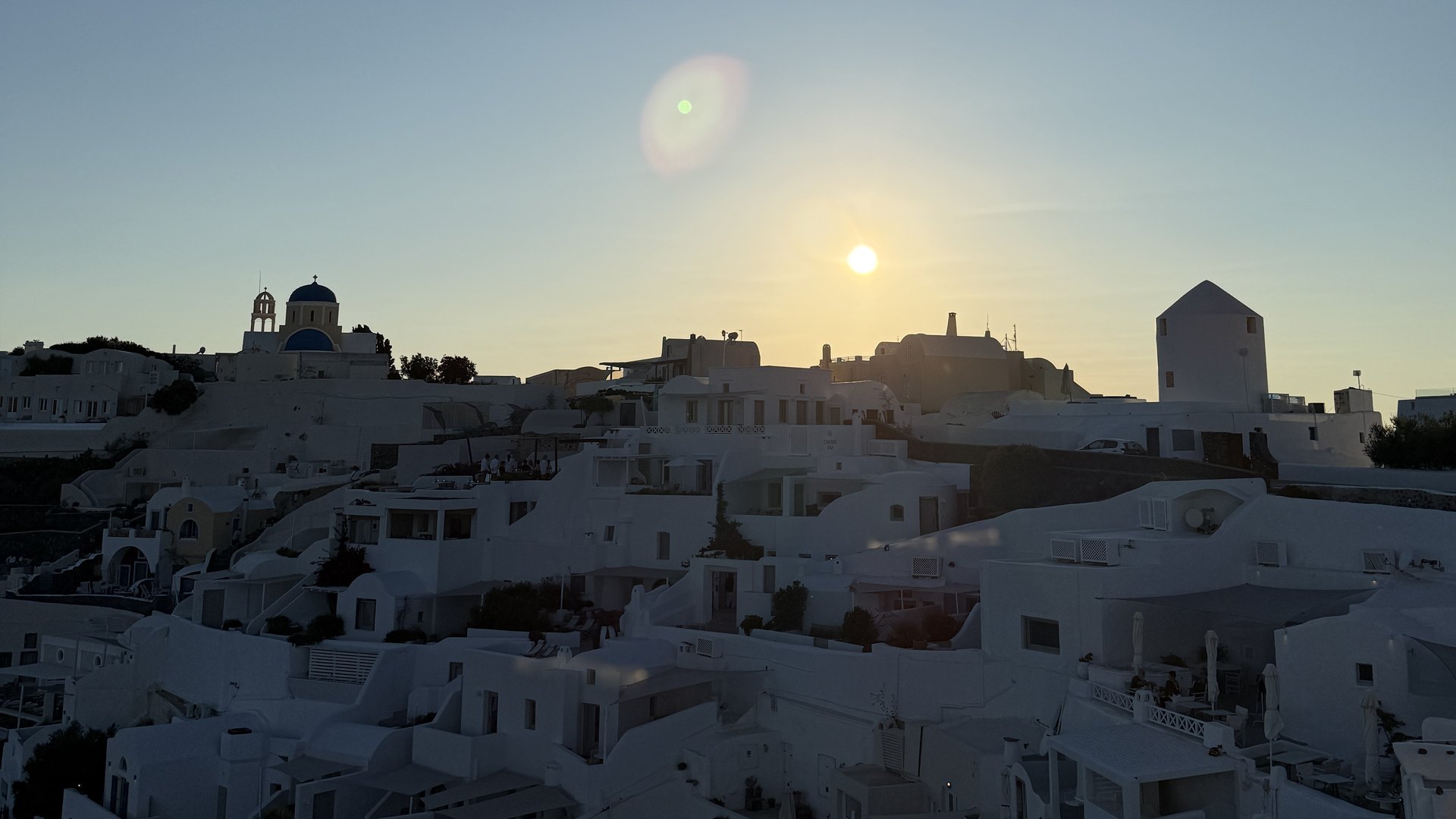 The sun setting over the white houses, blue dome and windmill of Oia