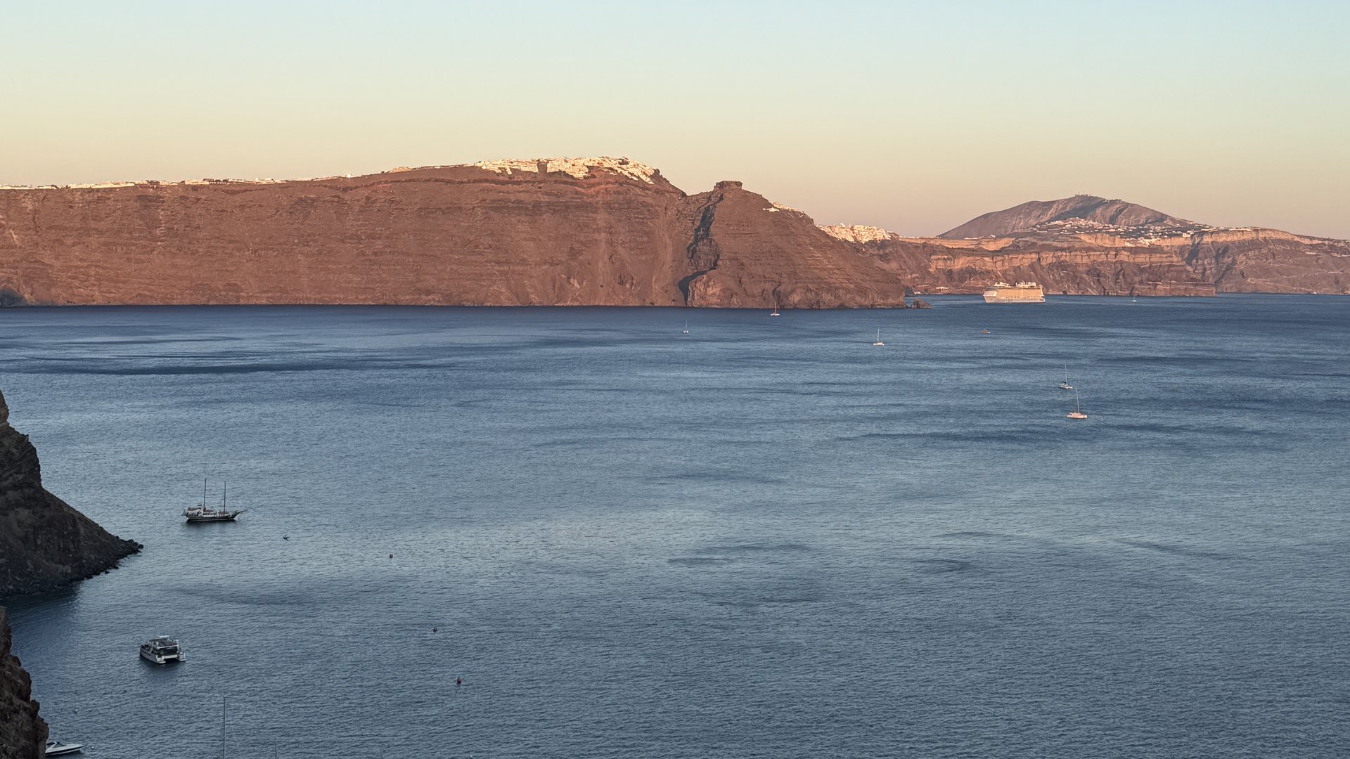 The caldera cliffs of Santorini lit orange at sunset, with boats on the water below