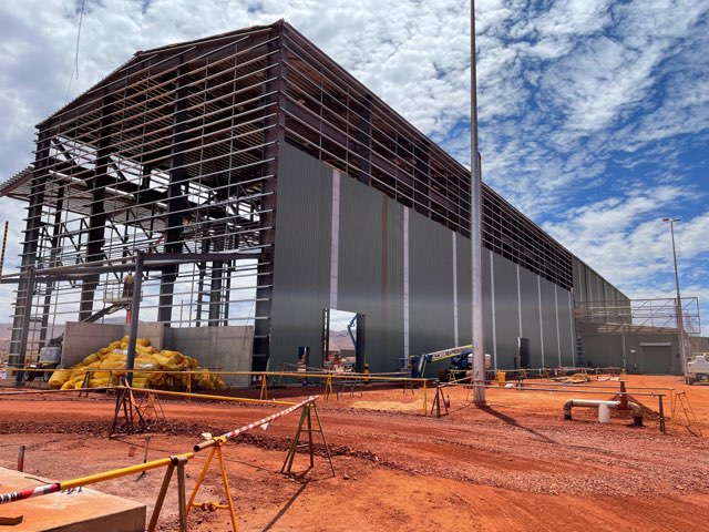 Steel-framed industrial shed being clad on a remote mining site with red dirt and safety barriers