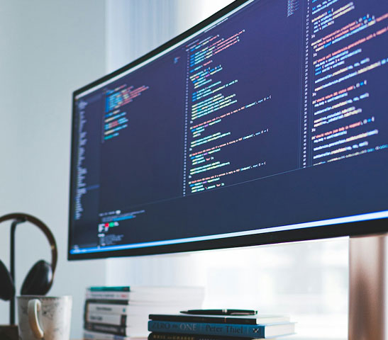 Ultrawide monitor displaying lines of code in a programming editor on a desk with books and headphones.