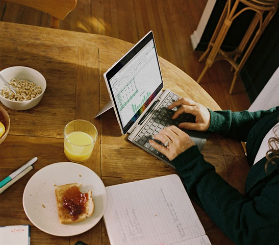 Person typing on a small laptop at a wooden table with toast, cereal bowl, and orange juice nearby.