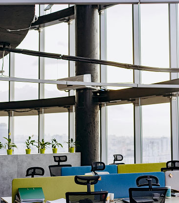 Modern office workspace with colorful chairs, desks, and potted plants by large floor-to-ceiling windows.