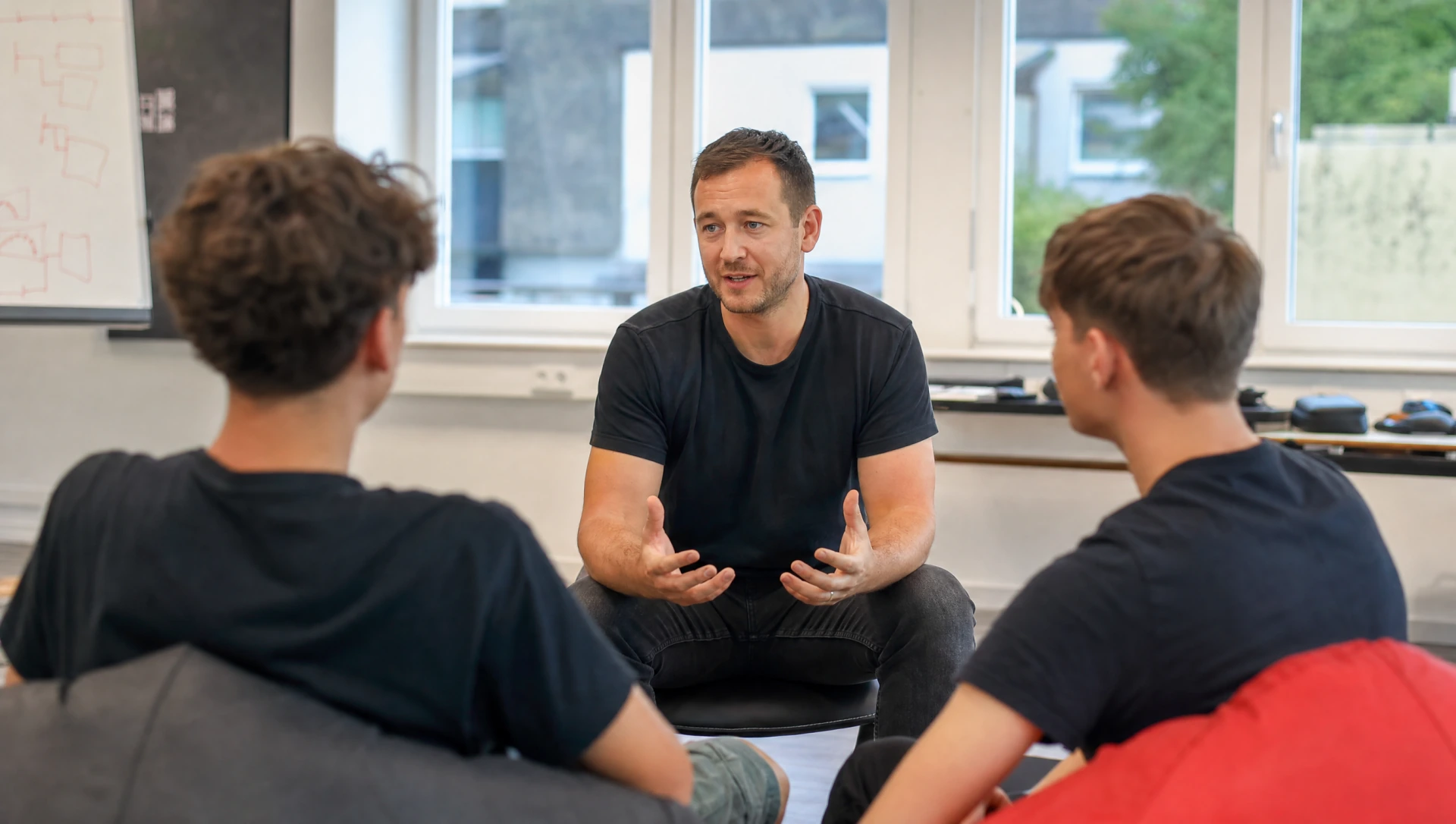 Trainer im schwarzen Shirt sitzt gestikulierend im Gespraech mit zwei Lehrlingen in offener Reflexionsrunde (KI-generiertes Bild)