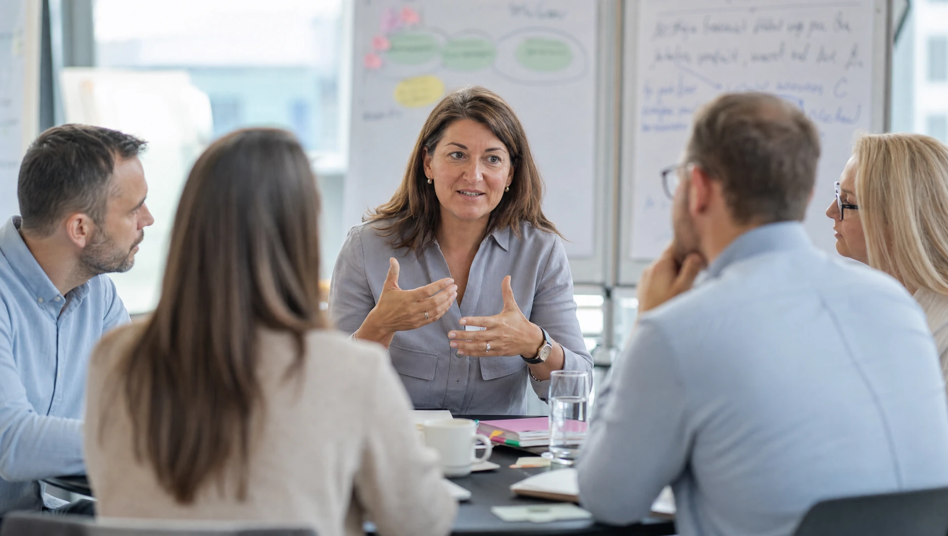 Moderatorin gestikuliert im Besprechungsraum, vier Teilnehmer:innen im Dialog vor Flipchart mit Mindmap (KI-generiertes Bild)