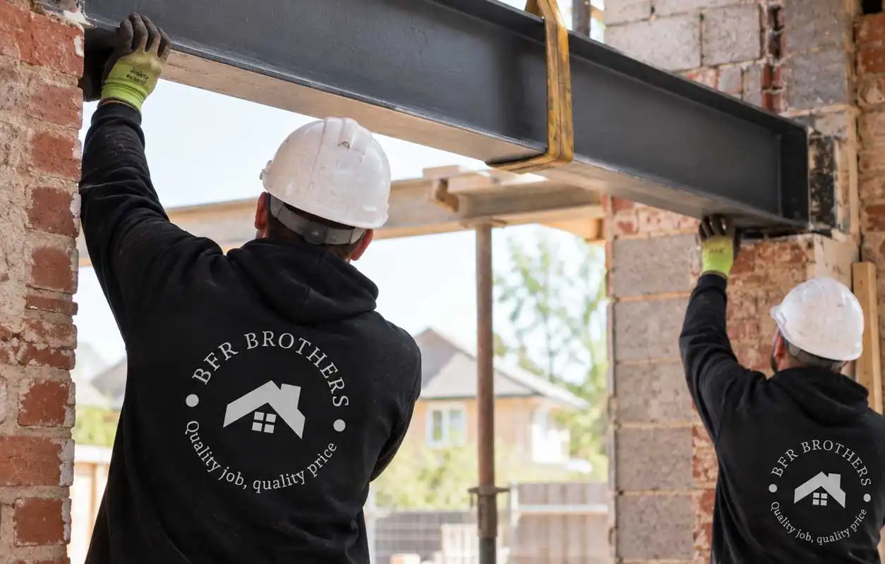 Two construction workers in hard hats install a steel I-beam into a brick wall opening using lifting straps, wearing black hoodies with BFR Brothers logo on site.