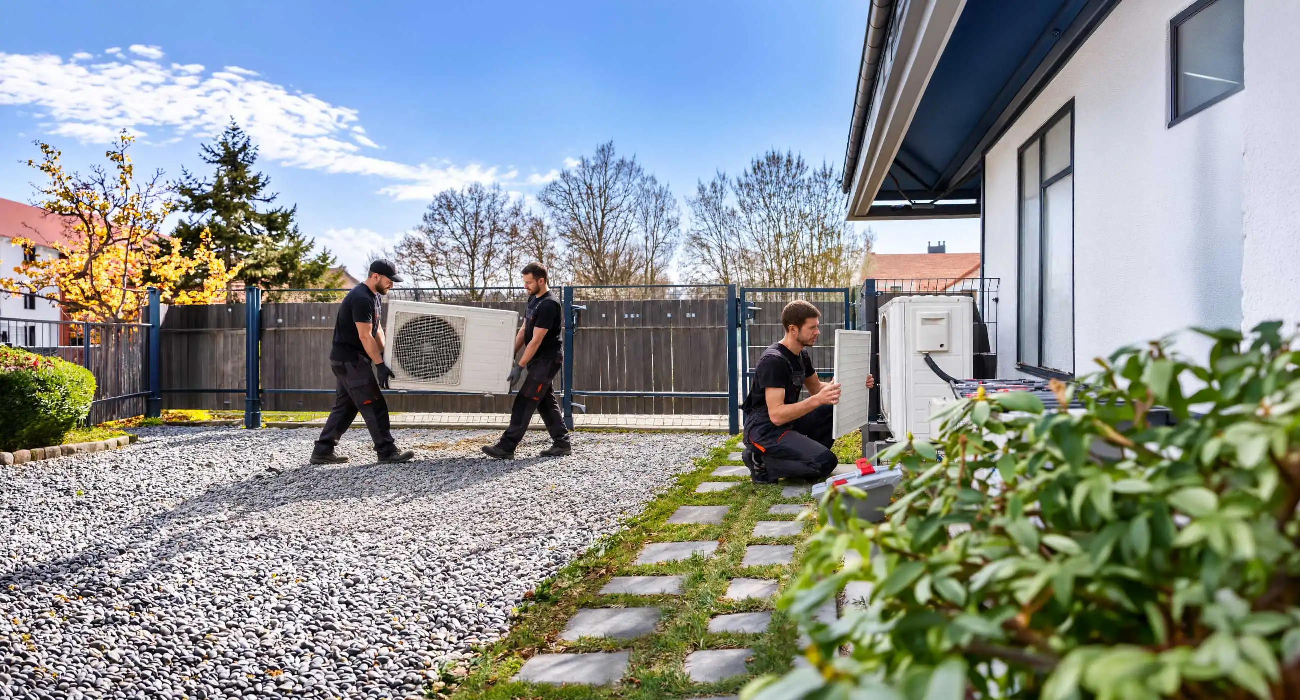 Technician servicing an outdoor heat pump unit, removing and inspecting the air filter beside a fenced residential property