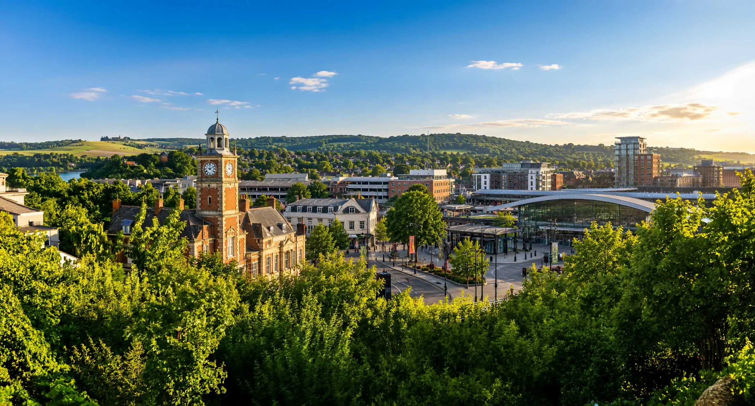 Panoramic view of Redhill town centre featuring modern station frontage, historic clock tower, surrounding buildings, and green hills under warm evening light in a wide 21:9 banner format