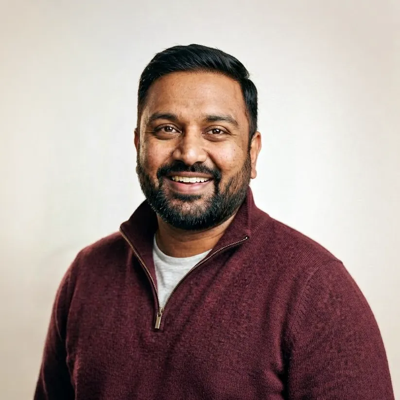 Smiling man with short black hair and beard wearing a maroon zip-up sweater over a white shirt against a plain light background.