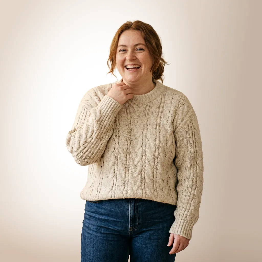 Smiling woman with red hair in a casual beige cable-knit sweater and blue jeans, standing against a light beige background.
