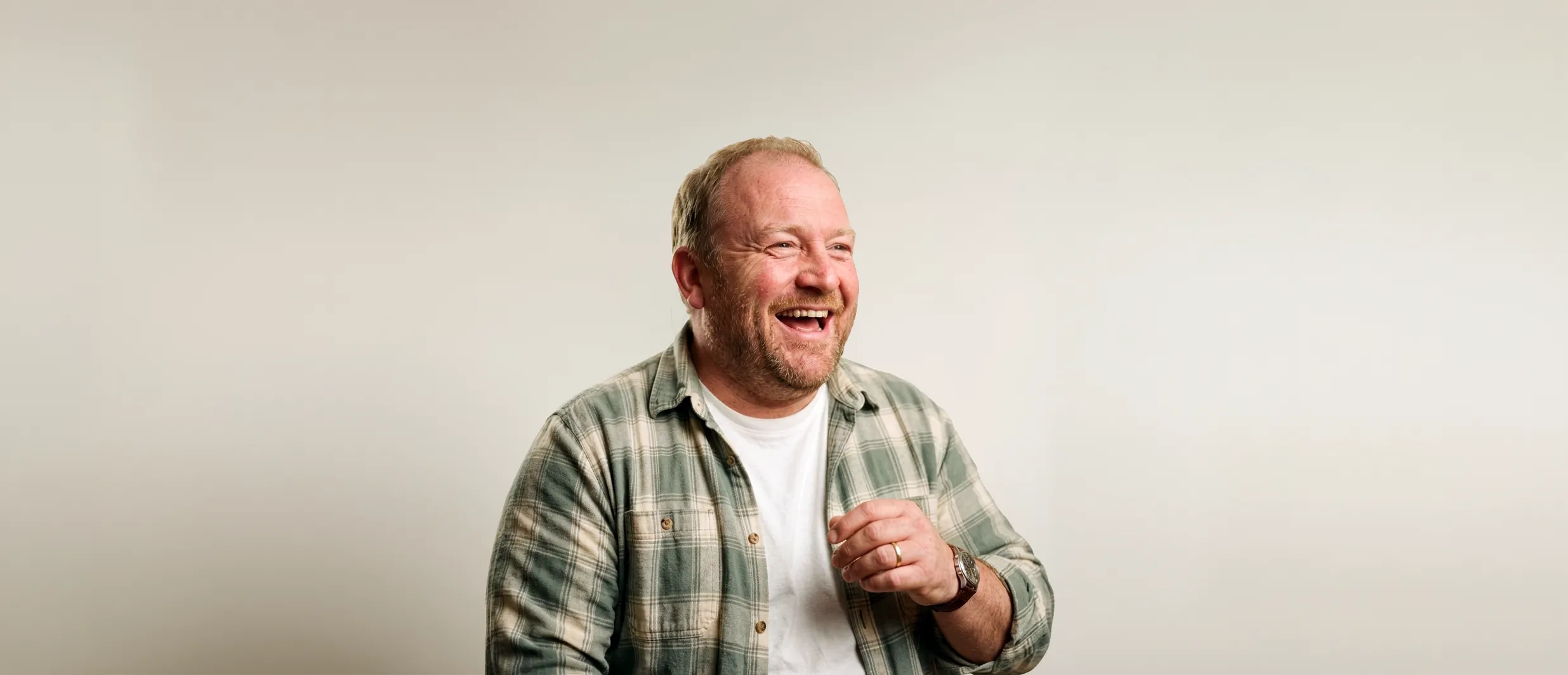 Smiling middle-aged man with a light beard wearing a green plaid shirt over a white t-shirt against a plain light background.