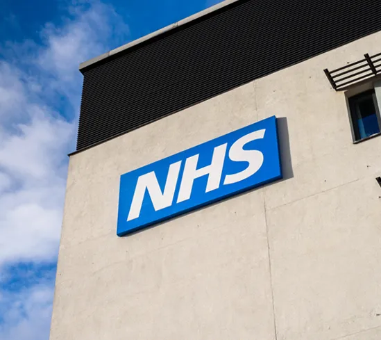Blue NHS sign with white letters mounted on a light-colored building exterior under a partly cloudy sky.