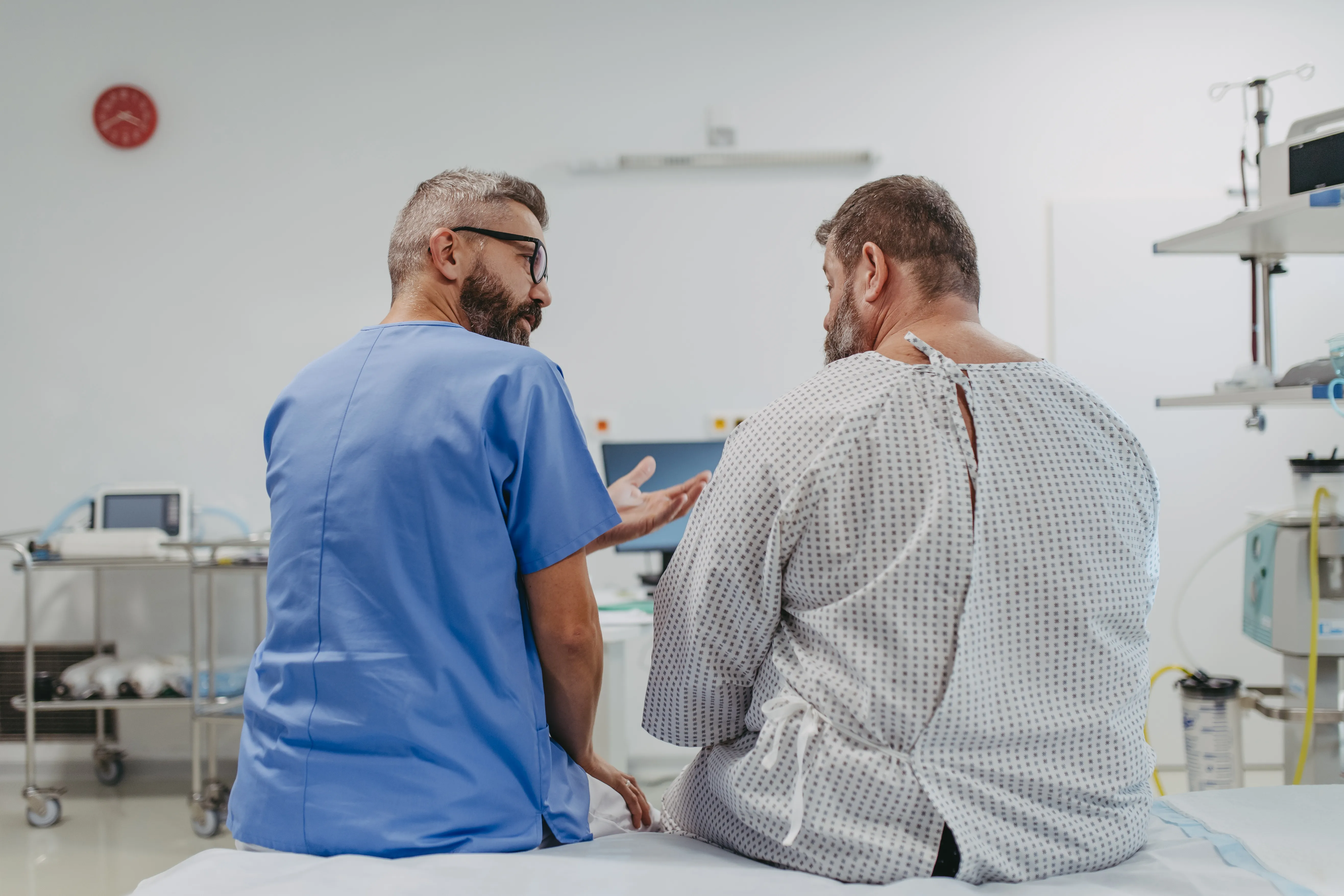 Male healthcare professional in blue scrubs talking to a male patient in a hospital gown sitting on an examination table in a clinical room.